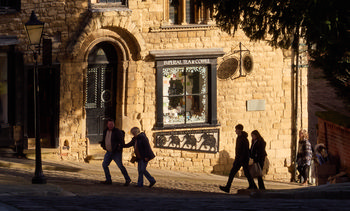 This urban photograph captures the famous Steep Hill in Lincoln, England, featuring the Imperial Tea shop as its main subject. Taken during the early afternoon in winter, the scene is bathed in low golden sunlight, casting long shadows typical of the season. Several people are seen walking along the cobbled street, wrapped in winter clothing, illustrating daily life in this historic part of Lincoln, United Kingdom. The honey-colored stone architecture and traditional shopfront emphasize the city's charm and heritage, making this area a notable landmark for locals and visitors alike. The image showcases a quintessential British setting with a focus on both the people and the unique character of this part of Lincoln.